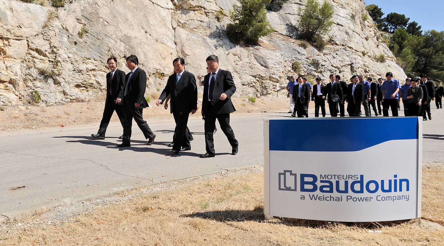 Group of people walking past a Moteurs Baudouin a Weichai Power Company sign.